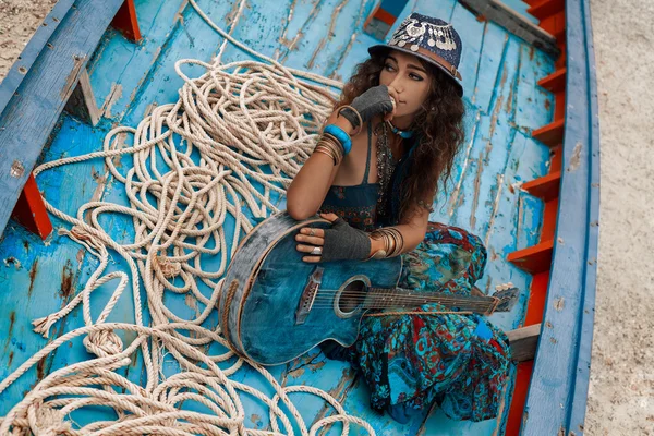 Στιβαρό και hippie νεαρή γυναίκα με κιθάρα που κάθονται σε παλιά βάρκα — Φωτογραφία Αρχείου