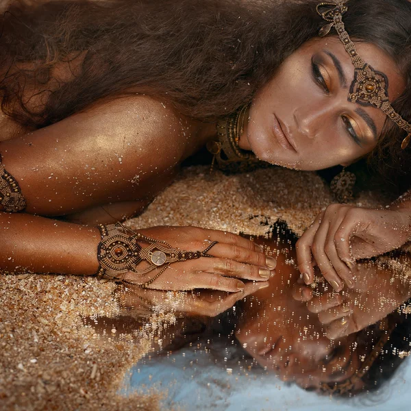 Mooi meisje in etnische juwelen en spiegel bedekt met zand — Stockfoto