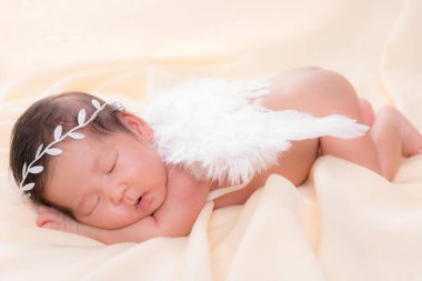 Bir aylık bir bebeğin portresi uyuyor, yeni doğmuş bir kız bebek. Beyaz bir saç bandı takıyor, melek kanatları takıyor ve krem rengi bir battaniyenin üzerinde uyuyor. Yeni doğan stüdyonun konsept portresi.