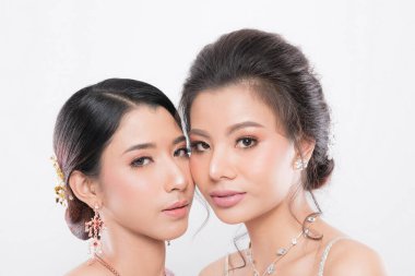 Yumuşak makyajlı ve karmaşık mücevherleri olan iki güzel Asyalı kadın, beyaz arka planda stüdyo çekimlerinde zarafeti sembolize ediyorlar..