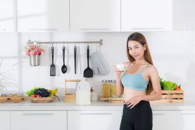 Mavi aktif giysili gülümseyen genç Asyalı kadın elinde parlak, modern bir mutfakta bir bardak taze süt tutarak sağlıklı beslenme alışkanlığını ve sağlığını vurguladı..