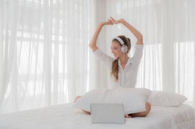 Beyaz tişörtlü, uzun saçlı, kulaklık takan neşeli beyaz kadın, beyaz yatakta dizüstü bilgisayarla oturup, parlak bir odada müzik eşliğinde kolunu kaldırıyor..