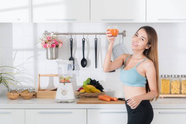 Temiz beyaz mutfakta mavi spor kıyafetli genç Asyalı bir kadın elinde renkli meyve püresi dolu bir bardakla kameraya neşeli bir ifadeyle bakar..