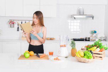 Mavi spor giysili sağlıklı genç Asyalı kadın bir yemek tarifi kitabına bakarken bir yandan da temiz beyaz mutfakta sağlıklı ve besleyici bir içecek hazırlayan bir blendere havuç ekliyor..