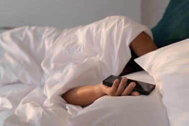 Sabaha kadar beyaz battaniyeli yatakta uyuyan bir adam akıllı telefon tutuyor.