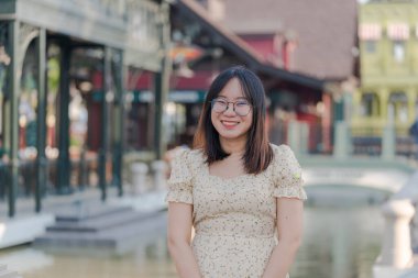 Renkli bir kasabada, nehire yakın mutlulukla gülümseyen gözlüklü Asyalı kadın portresi. Köprüde rahat rahat giyinen kadın.