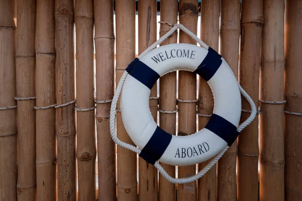 wooden bamboo wall with blue and white lifeguard ring hanging with text welcome aboard