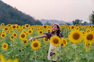 Şirin elbiseli mutlu bir Asyalı kadın ve Pele şapkalı. Çiçek açan sarı ayçiçeği bahçesinin keyfini çıkar. Kız çiçekli tatilde dinleniyor.