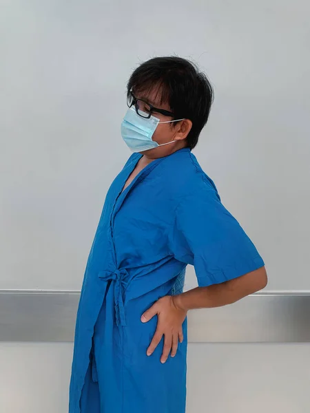 Asian man in blue hospital dress with protective face mask holding hand at side hip due to back pain, hip pain, medical illness