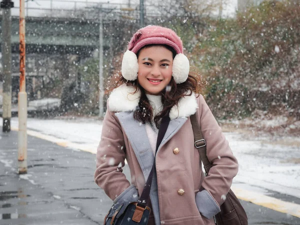 幸せなアジアの女性で冬の服でベレー帽とふわふわの耳フ立って鉄道駅プラットフォームで雪のシャワー 女の子は冬の休暇にリラックス — ストック写真