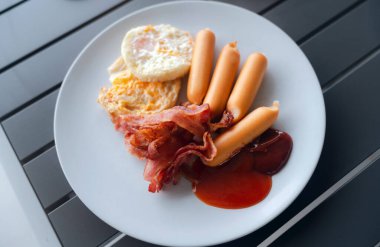 Beyaz tabakta ev yapımı Amerikan kahvaltısı, kızarmış pastırma, sosis, sahanda yumurta ve domates sosu ahşap masada..