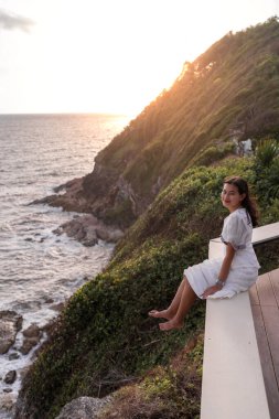 Asyalı kadın turist, akşamları uçurumların ve denizin üzerindeki teras koltuğunda oturuyor. Phasuk Nirun, Ko Proet, Chanthaburi, Tayland 'da güneş ışığı, gökyüzü manzaralı.