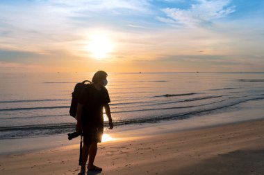 Siluet Asyalı, koruyucu yüz maskesi takan sırt çantalı adam Huahin, Tayland 'da sıcak bir sabah güneşiyle sahilde yürüyen kamerayı tut.