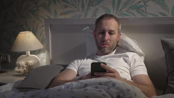 寝る前にスマートフォンでベッドに横たわっ若い男. — ストック動画