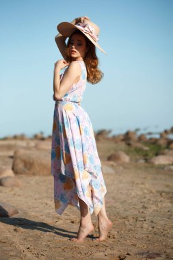 Geniş kenarlı hasır şapkalı ve yaz elbisesiyle, sıcak bir yaz gününde plaj arka planında sevimli bir kız..