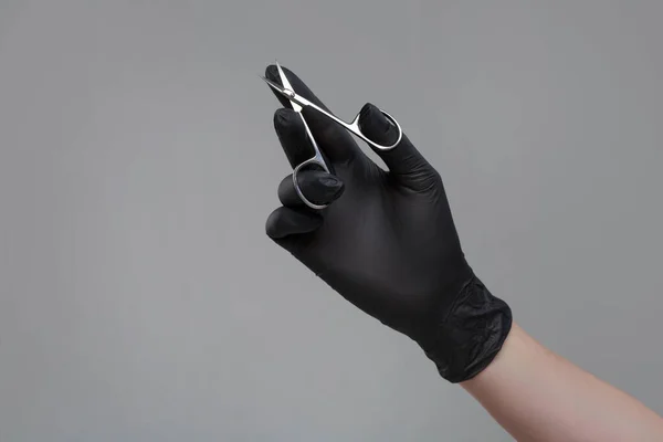 Female hand in a black latex glove holds nail scissors Isolated on a gray background.