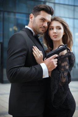 Zarif, zarif bir adam elinde silahla dışarıda güzel bir kıza sarılıyor..