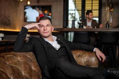 Koyu renk üç parçalı takım elbiseli zarif genç bir adam bir restoranın dinlenme yerinde deri bir koltukta oturuyor..