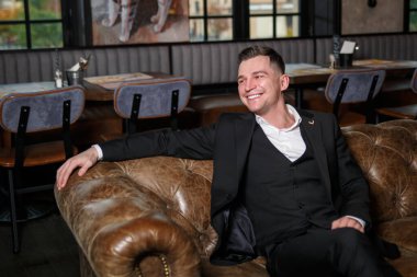 Koyu renk üç parçalı takım elbiseli zarif genç bir adam bir restoranın dinlenme yerinde deri bir koltukta oturuyor..