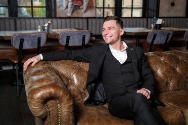 Koyu renk üç parçalı takım elbiseli zarif genç bir adam bir restoranın dinlenme yerinde deri bir koltukta oturuyor..