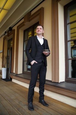 Üç parçalı şık takım elbiseli çekici genç bir adam elinde bir fincan kahveyle terasta duruyor..