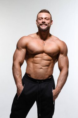 Çıplak gövdeli, sırıtan genç kaslı bir adam açık gri bir arka planda tek başına duruyor..
