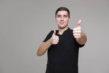 Siyah iş kıyafetleri içinde genç beyaz bir adam iki elinde baş parmağını gösteriyor, gri arka planda izole edilmiş..