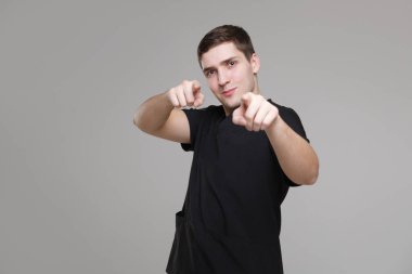 Gündelik çalışan siyah elbiseli genç beyaz adam gri bir arka planda iki elini de işaret ediyor..