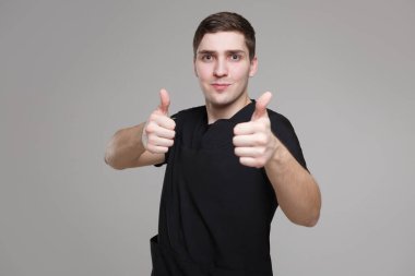 Siyah iş kıyafetleri içinde genç beyaz bir adam iki elinde baş parmağını gösteriyor, gri arka planda izole edilmiş..