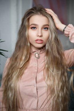 Güzel sarışın, uzun saçlı, narin, eski moda bir kız. İç kısımlarında toz pembe bir elbise giyiyor..