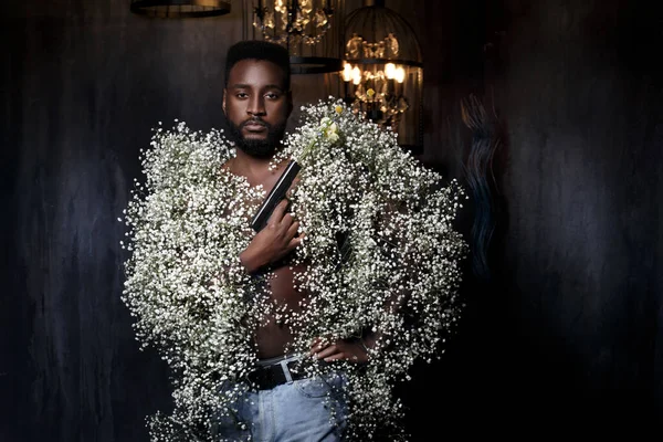 Fashion portrait of a black man in a gypsophila coat holding a pistol shooting a bouquet of flowers.