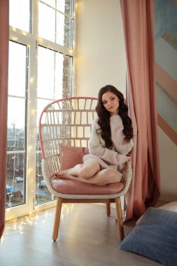 Hoş, genç, esmer, rahat kazaklı bir kız pencerenin yanındaki sandalyede oturuyor..