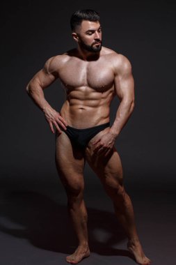 Sakallı genç atletik erkek sporcu koyu bir arka planda tamamen büyüyen kaslar gösteriyor..