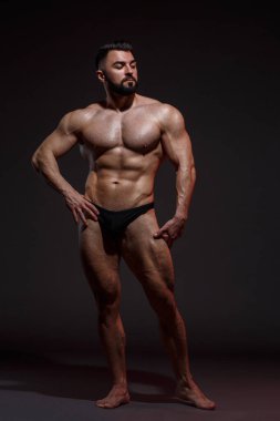 Sakallı genç atletik erkek sporcu koyu bir arka planda tamamen büyüyen kaslar gösteriyor..