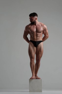 Atletik zayıf adam, gri arka planda izole edilmiş bir heykel gibi, bir küp üzerinde tam gelişim içinde duruyor..