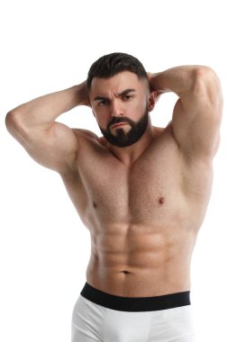 Çıplak kaslı sakallı, atletik bir adam beyaz arka planda iki elini de başının arkasında tutuyor..