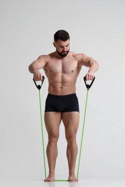 Genç atletik sakallı adam, gri arka planda izole edilmiş elastik bantla spor yapmaya gider..