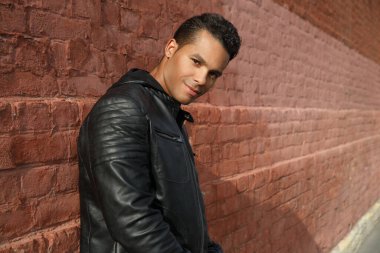 Siyah deri ceketli yakışıklı bir adam gülümsüyor ve dışarıdaki kırmızı tuğla duvara yaslanıyor..