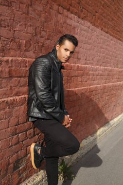 Siyah deri ceketli yakışıklı adam, dışarıdaki kırmızı tuğladan duvara yaslanıyor..