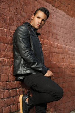 Siyah deri ceketli yakışıklı adam, dışarıdaki kırmızı tuğladan duvara yaslanıyor..