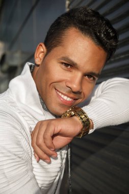 Yüzünde bir gülümseme olan, dışarıda siyah bir arka planda şık beyaz günlük kıyafetler giyen, mutlu, çekici bir Latin adamın portresi..