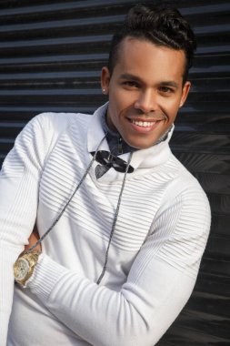 Yüzünde bir gülümseme olan, dışarıda siyah bir arka planda şık beyaz günlük kıyafetler giyen, mutlu, çekici bir Latin adamın portresi..