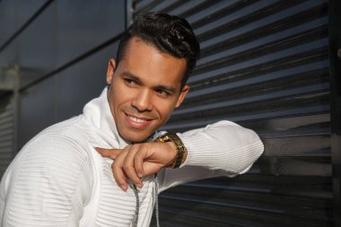 Yüzünde bir gülümseme olan, dışarıda siyah bir arka planda şık beyaz günlük kıyafetler giyen, mutlu, çekici bir Latin adamın portresi..