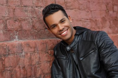 Siyah deri ceketli genç, mutlu Latin adam kırmızı tuğla bir duvarın arkasında gülümsüyor..
