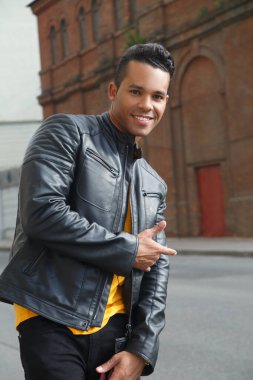 Genç, çekici, gülümseyen bir Latin adamın portresi, elleri sağda, bir şehir caddesinde sergileniyor..