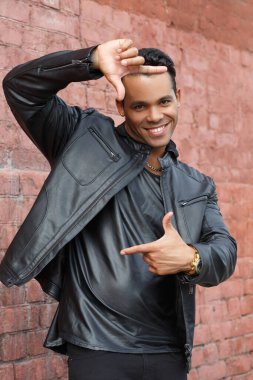 Siyah deri ceketli, mutlu, genç Latin bir adam elleri ile kırmızı tuğlalı bir duvarın arkasında bir çerçeve şeklinde gülümsüyor..