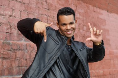 Siyah deri ceketli, mutlu, genç Latin bir adam elleri ile kırmızı tuğlalı bir duvarın arkasında bir çerçeve şeklinde gülümsüyor..