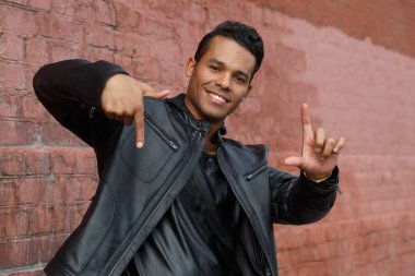 Siyah deri ceketli, mutlu, genç Latin bir adam elleri ile kırmızı tuğlalı bir duvarın arkasında bir çerçeve şeklinde gülümsüyor..