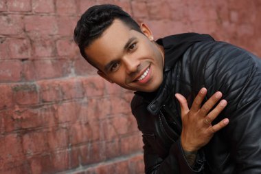 Siyah deri ceketli ve gülümseyen genç, mutlu, Latin bir adam kalbini kırmızı tuğla bir duvarın arka planına dayadı..