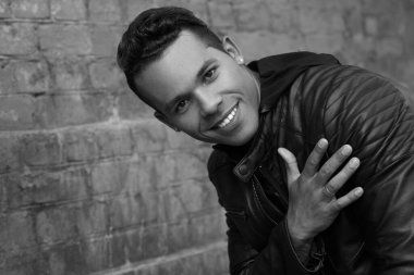 Siyah beyaz bir portre, siyah deri ceketli, mutlu, genç bir Latin adam gülümsüyor ve kalbini avucuyla kırmızı tuğla bir duvarın arkasına dayadı..